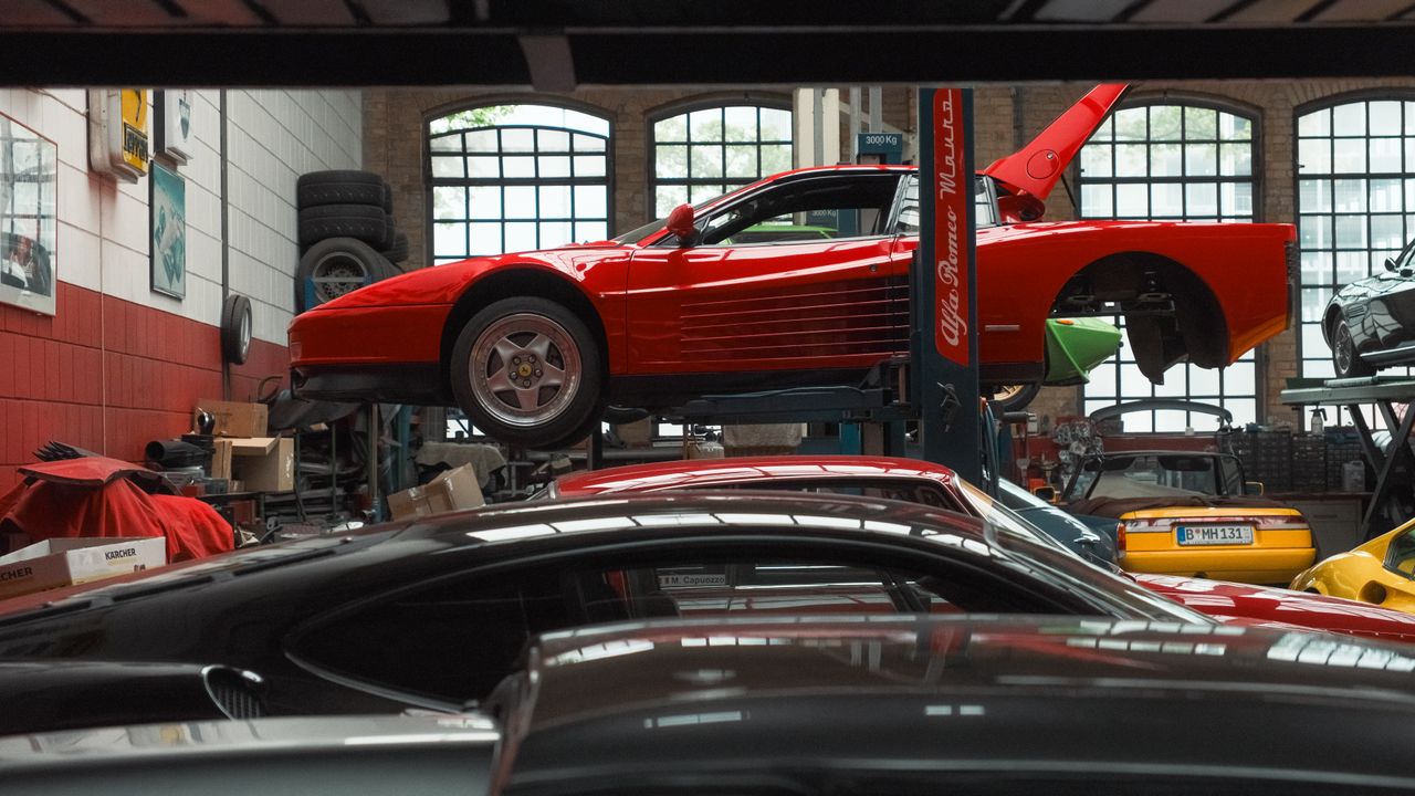 services-01 A vibrant red sports car is elevated on a lift inside an auto repair garage, surrounded by various vehicles and equipment.